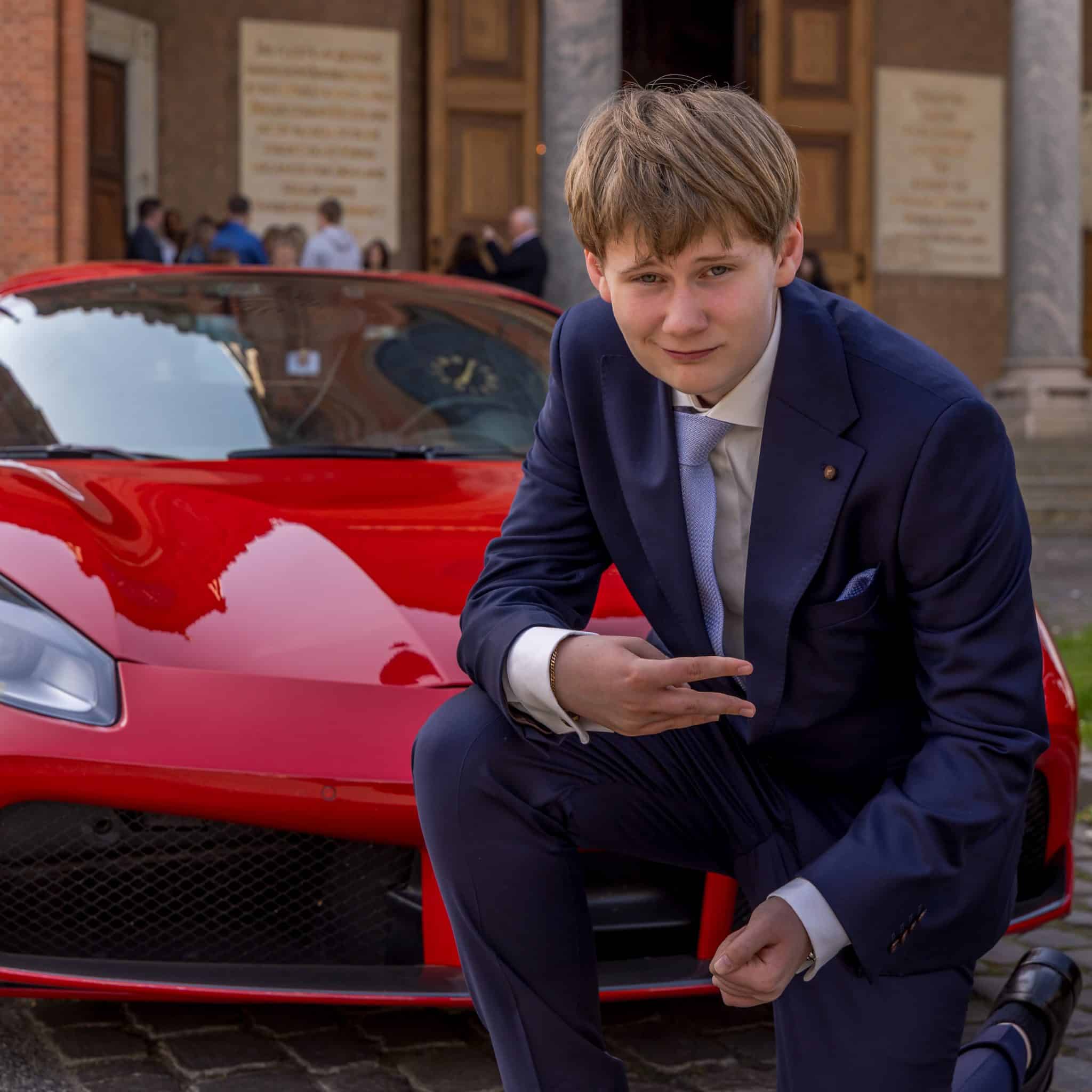 Konfirmationsfoto af dreng i marineblå suit foran rød Ferrari sportsvogn ved kirke – professionelt konfirmationsfoto