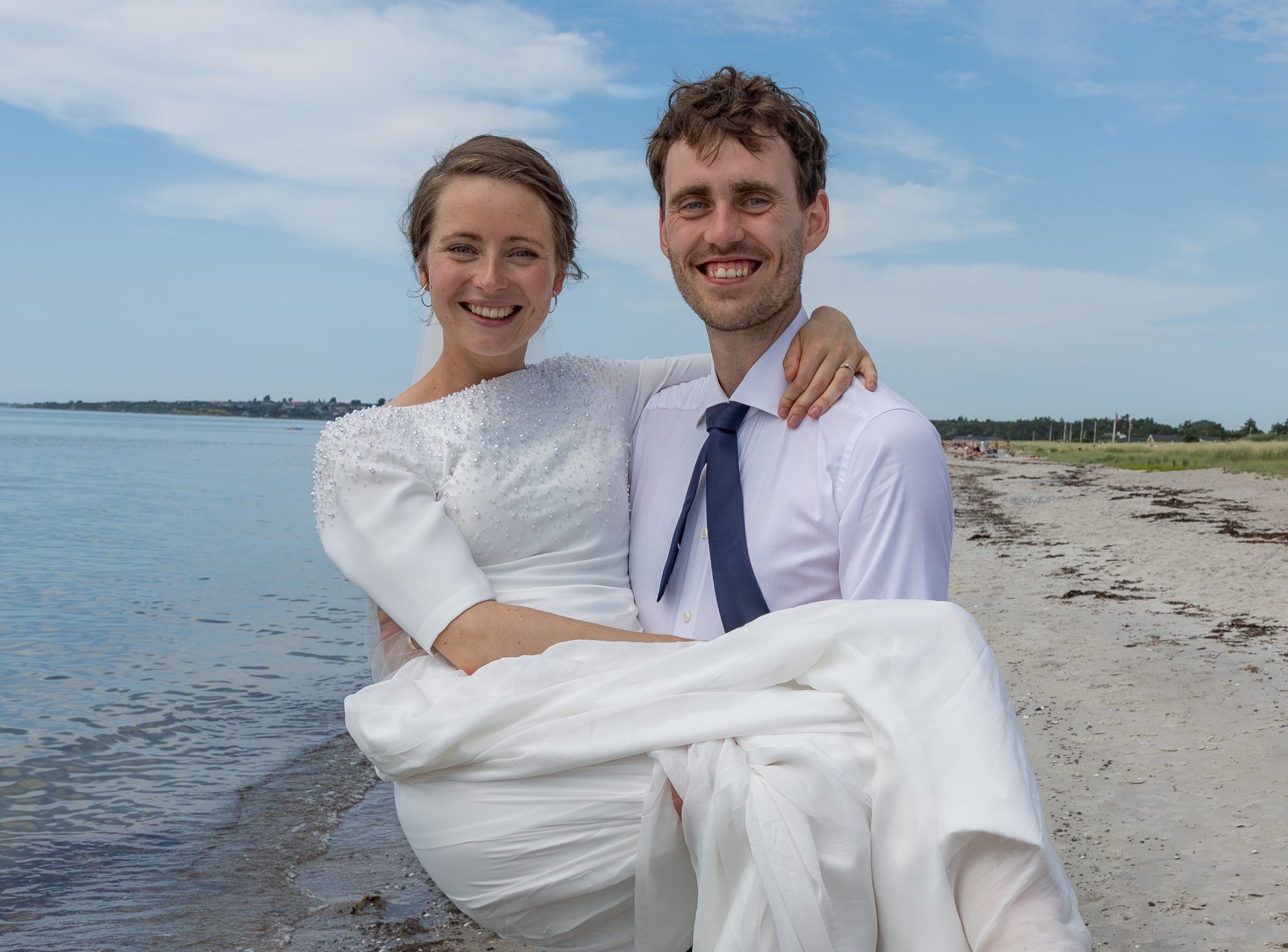 Festfotograferne har her fotograferet brudeparret sammen på stranden med havet og den blå himmel i baggrunden. Gommen bærer bruden.