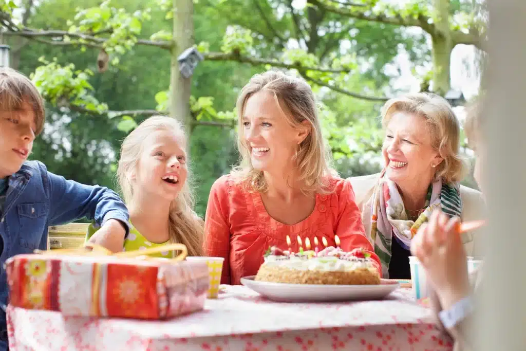 Festfotograferne har her fotograferet til en familiefødselsdag i haven. Her ses et borde med en gave og en kage med lys i. Ved bordet sidder 2 børn (en dreng og en pige) en kvinde i en orange bluse og en lidt ældre kvinde ved siden af.