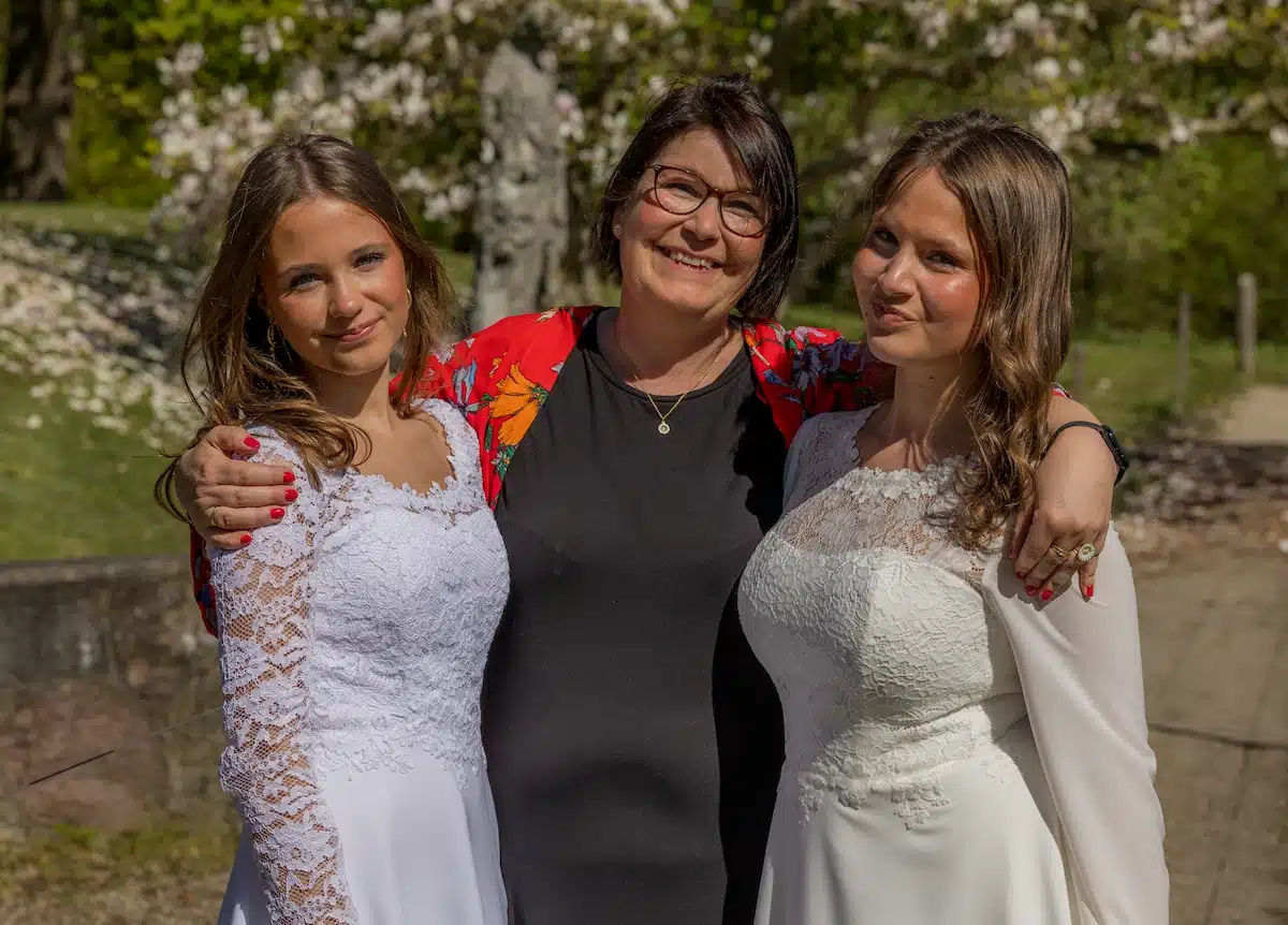 Her har festfotograferne fotograferet 2 tvillingepiger på deres konfirmationsdag. På billedet står de i en have med deres mor mellem sig, der holder begge piger rundt om deres skuldre. I baggrunden ses et blomstrende hvidt træ