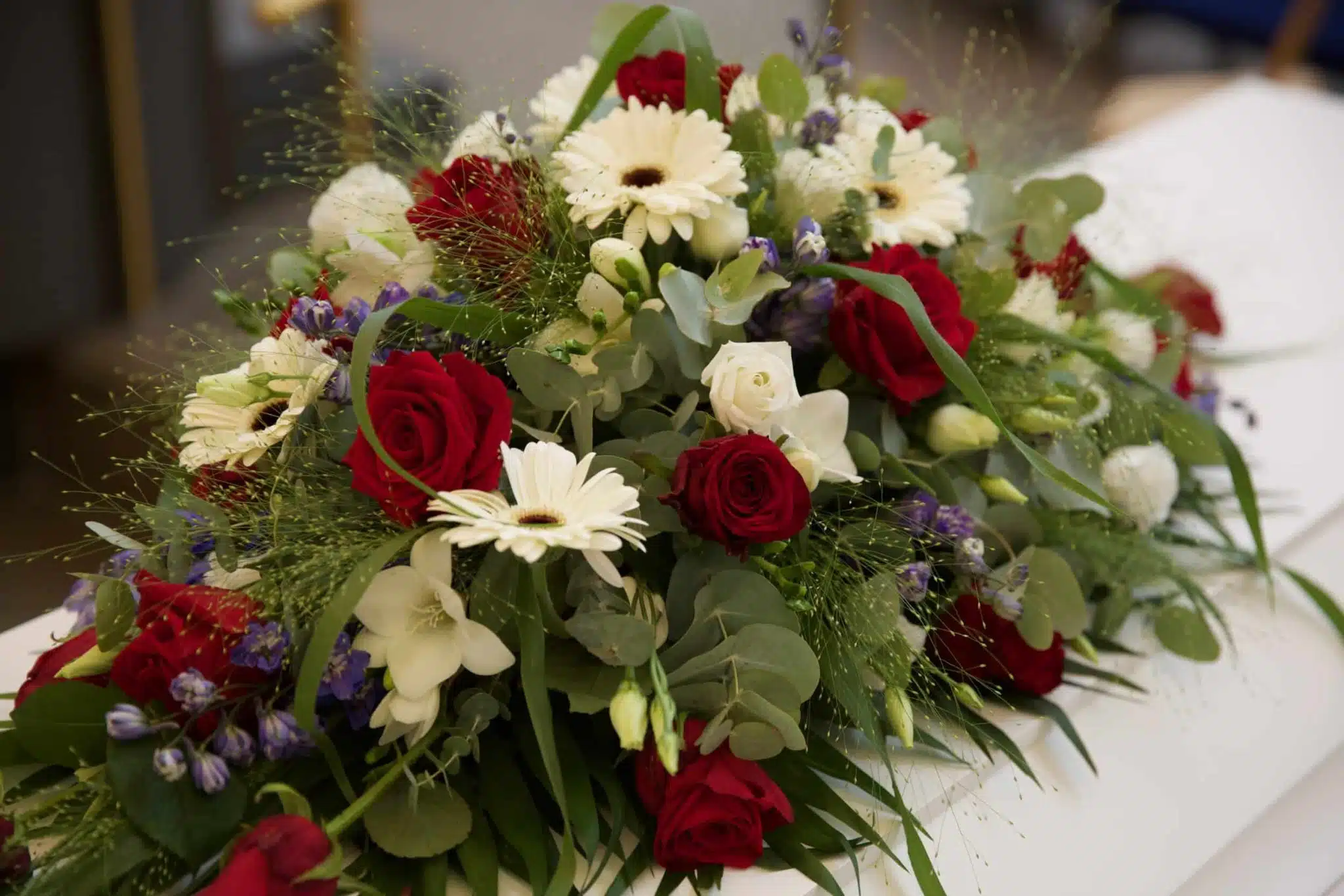 På billedet ses en bårebuket i hvide,lilla og røde nuancer på en kiste. Bårebuketten fylder hele billedet