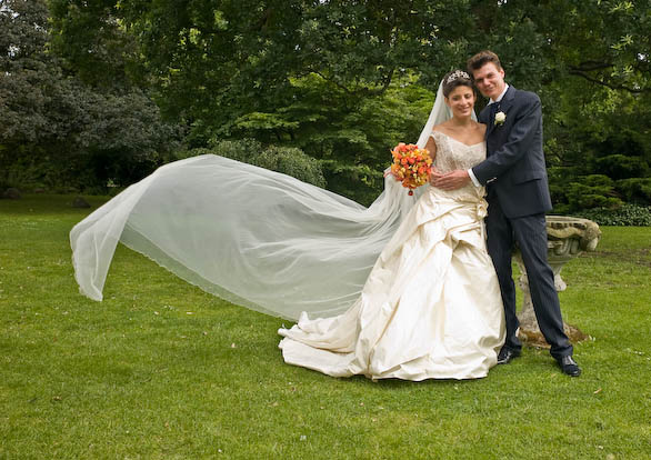Festfotograferne har her fotograferet et Brudepar opstillet i have. Brudens slør flagrer i vinden. Hun bærer desuden en brudebuket i orange nuancer