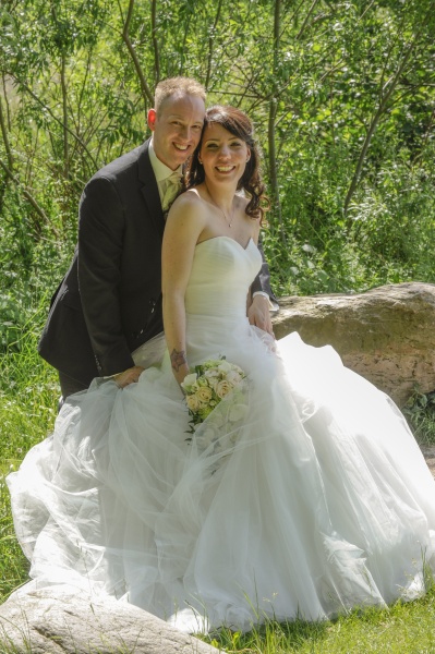 festfotograferne har her fotograferet et brudepar i en have. Bruden ses siddende på en sten med sin store flotte kjole og holdende sin brudebuket i hvide farver. Gommen står bag hende foroverbøjet, så hans kind støder mod hendes tinding