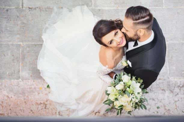 Festfotografen har her fotograferet bryllupsbillede. Billedet er taget oppefra. Bruden kigger op i kameraet og holder sin brudebuket i hvide nuancer, mens brudgommen kysser bruden på kinden