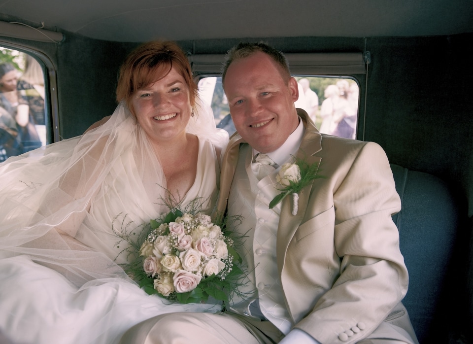 Festfotograferne har her fotograferet et brudepar, der sidder i en bil. Bruden, der er rødhåret, bærer slør og sidder med sin brudebuket i hvide nuancer. Gommen er i beige jakkesæt med hvid skjorte og en hvid rose i kanppehullet