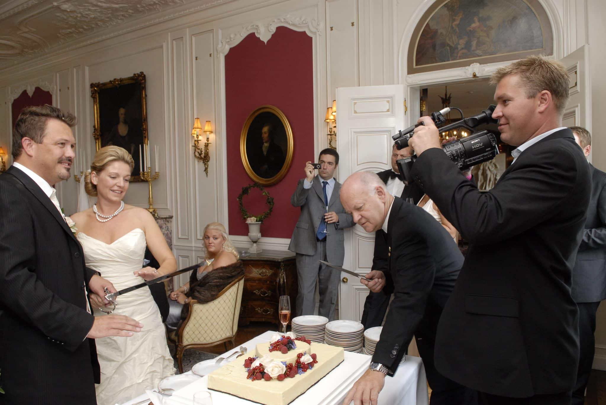Festfotograferne har her filmet og fotograferet til en bryllup. På billedet ses et brudepar, der skal til at skære for i bryllupskagen med en sabel ved en reception. Festfotografens videograf ses ligeledes i billedet med sit videokamera