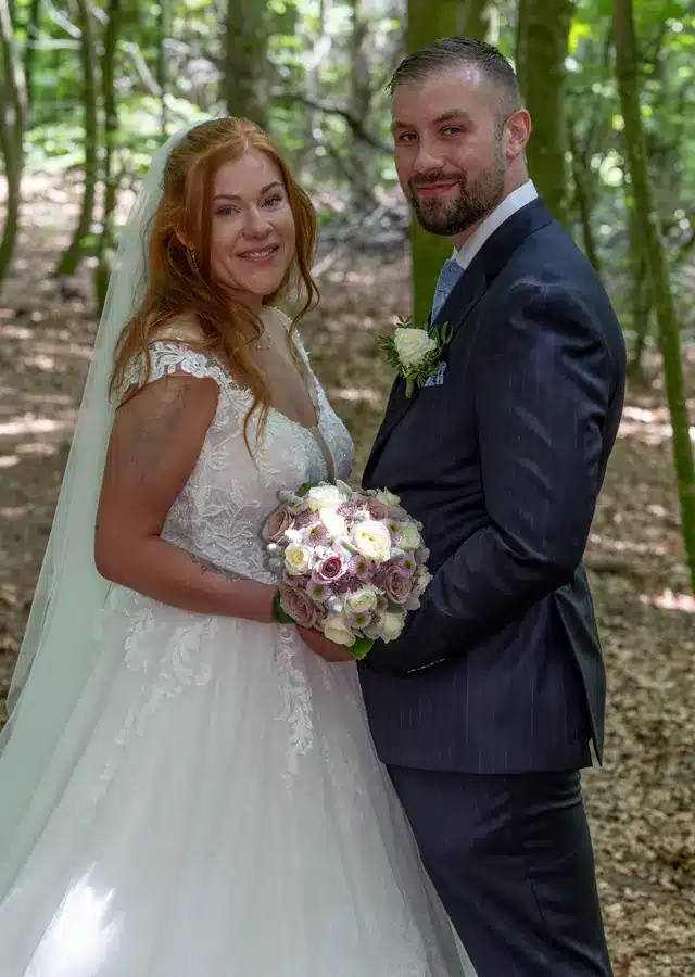 Bryllup 18 Brudepar fotograferet i skov. Parret står og holder om hinanden og brudebuketten, mens de kigger i kameraet