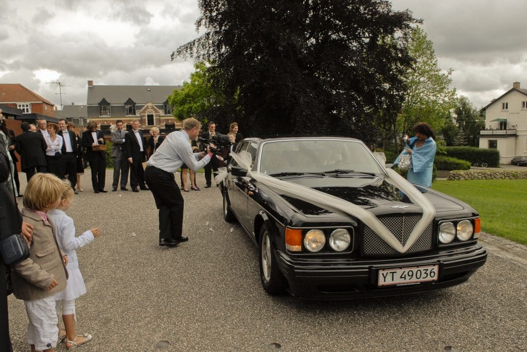 Festfotografen har her fotograferet til et bryllup. På billedet ses bilen med brudeparret køre væk, mens gæsterne står og ser den køre væk. Festfotografernes fotograf står ved siden af bilen og filmer ind i bilen