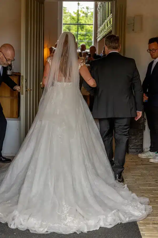 Brud i hvid kjole med slæb følges ind i kirke - bryllupsfoto kirkegang