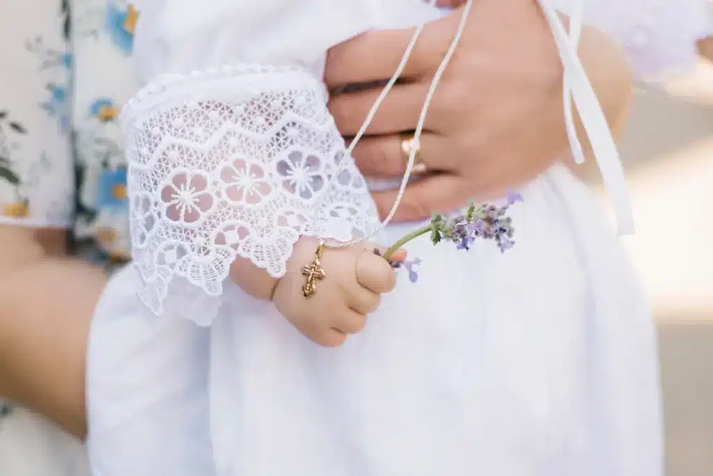 Billede af hånden på et barn der skal døbes. Hun holder en lavendel blomst i hånden. Barnet bliver holdt af en voksen