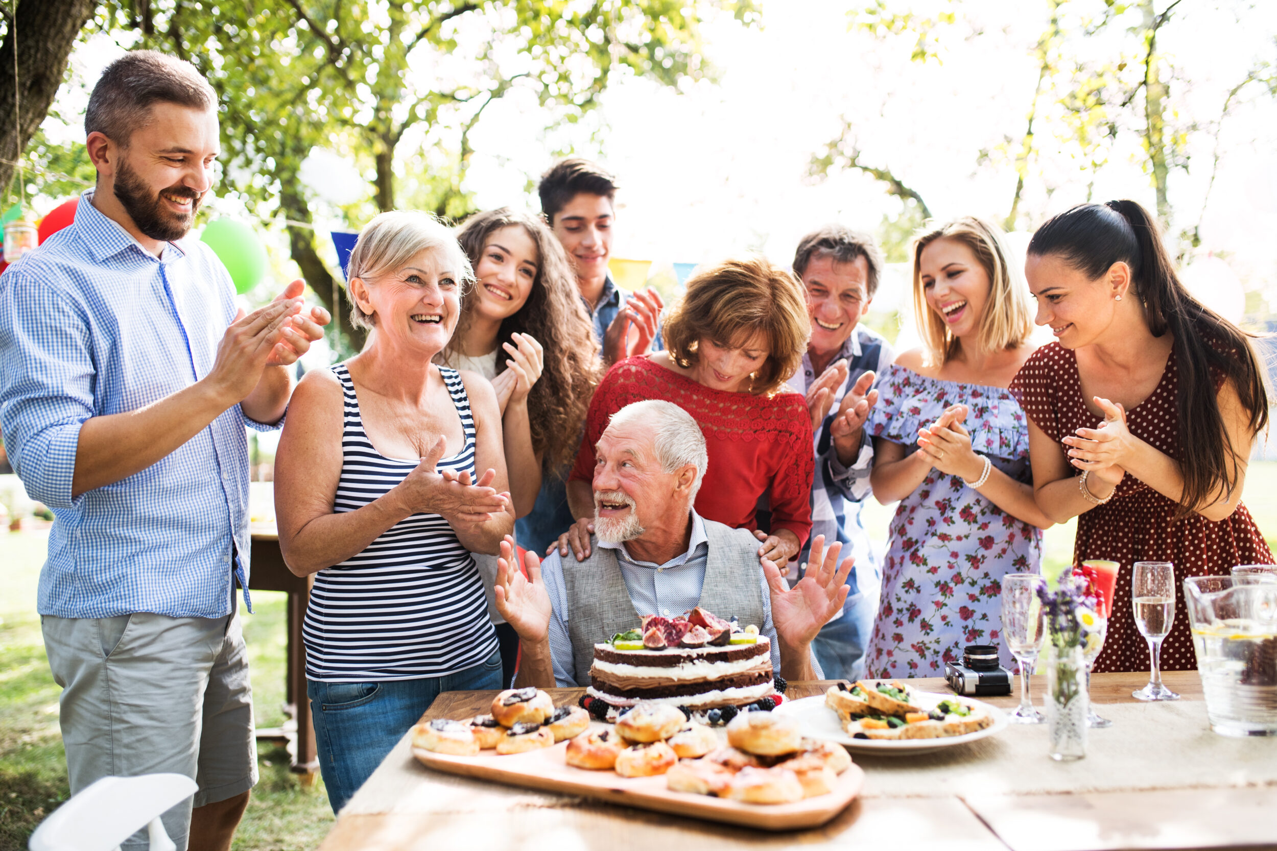 Familie står omkring siddende bedstefar. Han sidder ved bordet, der er fyldt med kager og champagne