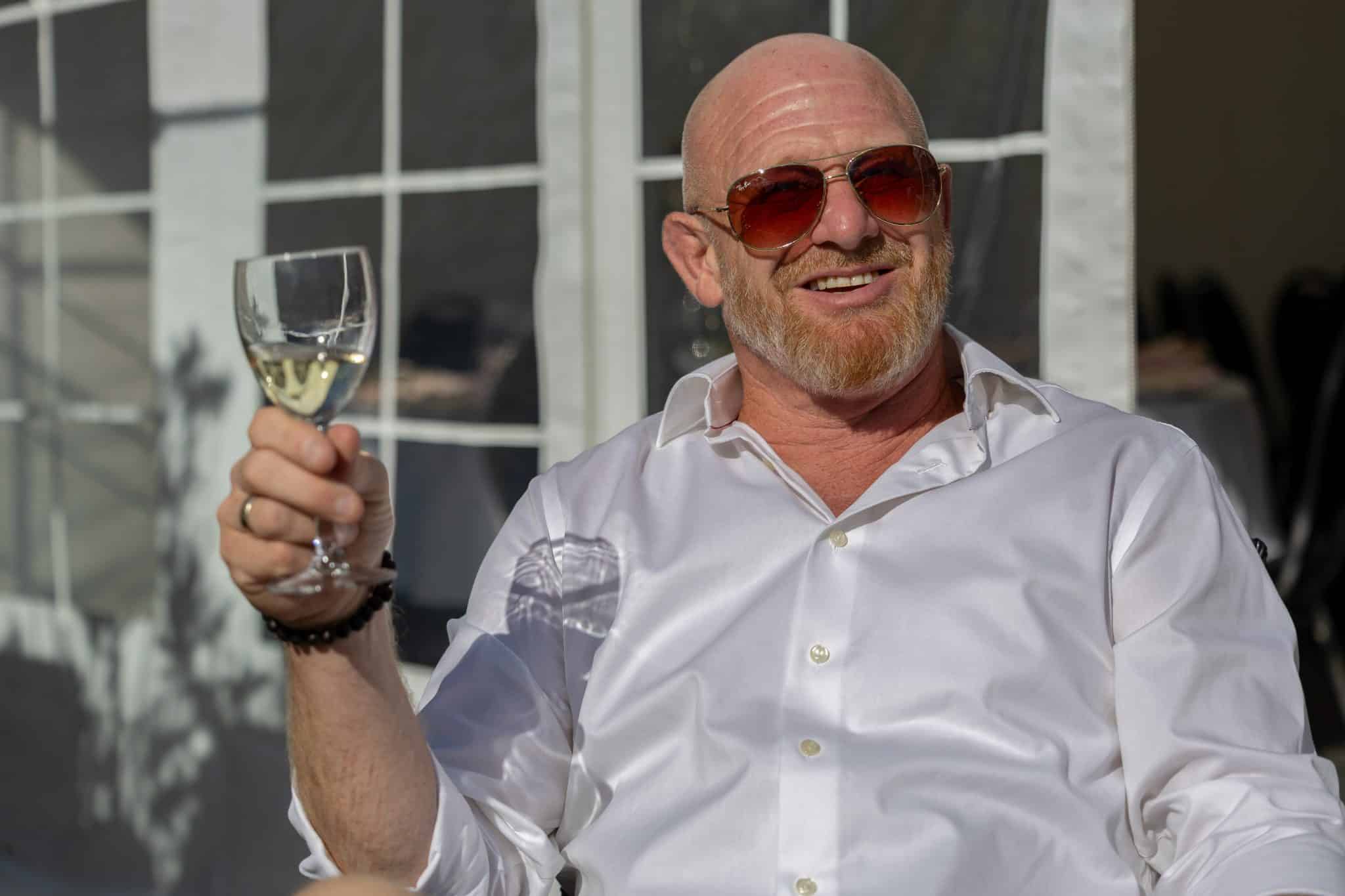 Skaldet mand med solbriller sidder med vinglas i hånden og nyder solen