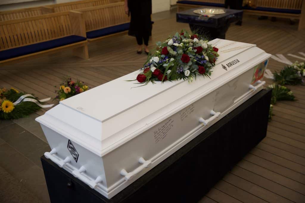 På billedet ses en hvid kiste med en bårebuket i hvide, lilla og røde nuancer. På kisten er der skrevet hilsner til afdøde