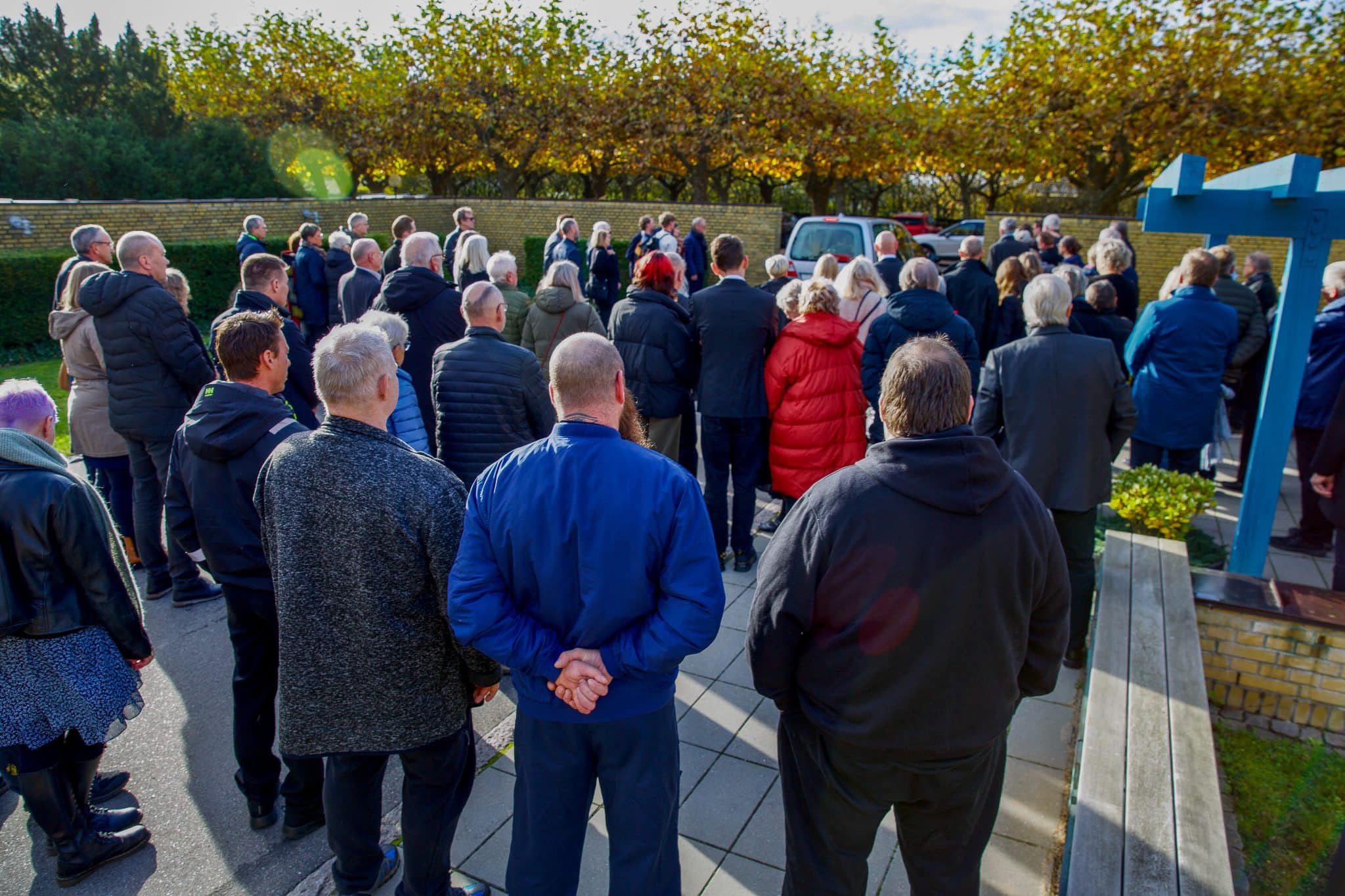 Flok af mennesker til begravelse, der står med ryggen til kameraet, står og ser på at rustvognen kører væk