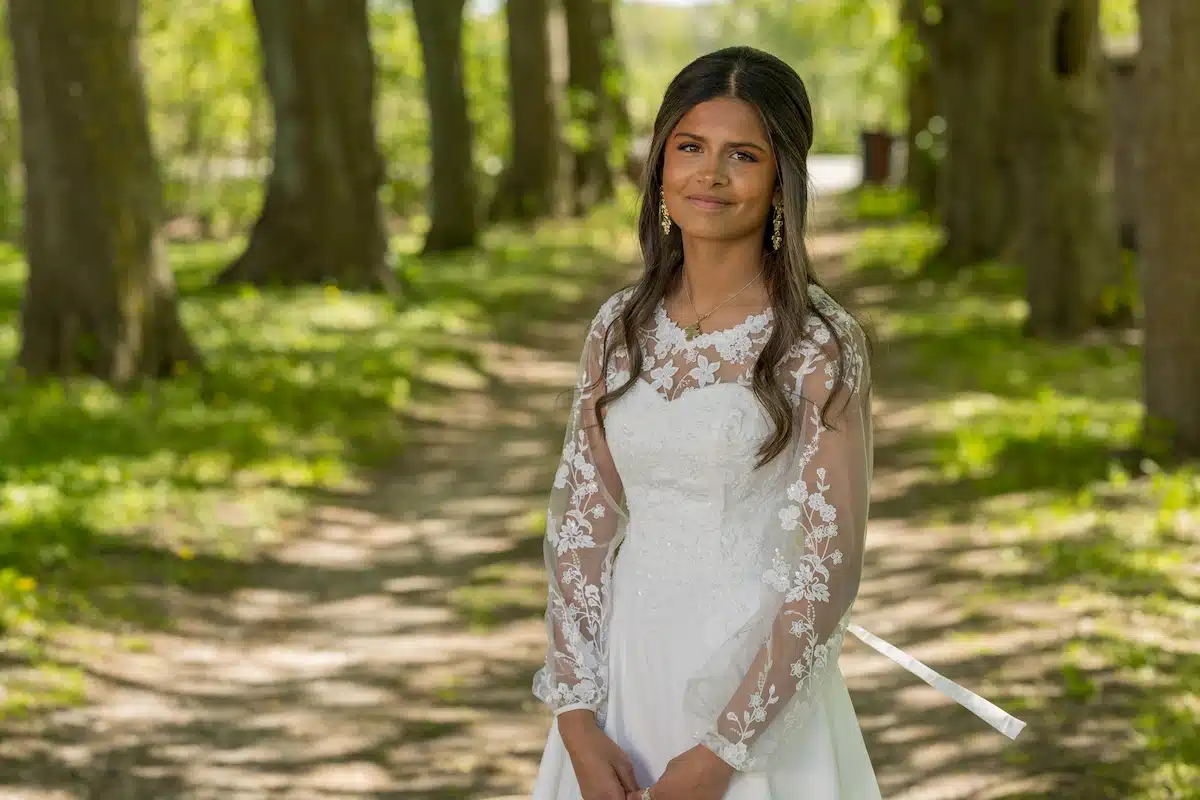 Konfirmationsfoto af pige i hvid blondekjole med blomstermønster og guldøreringe – professionelt konfirmationsfoto i smuk skovallé med solskin