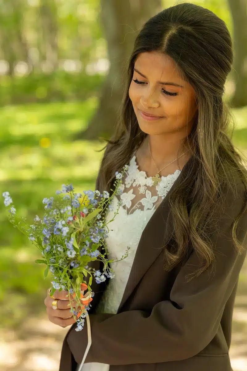 Konfirmationsfoto af pige i hvid blondekjole og brun blazer med buket blå forårsblomster – professionelt konfirmationsfoto i skov