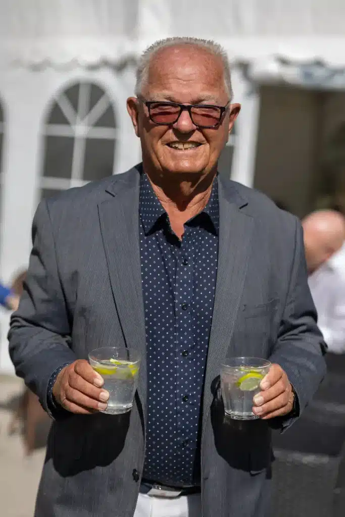 Festfotograferne har her taget et billede af en mand til havefest. Han står i grå blazer og en blå skjorte. Han har to drinks i hænderne