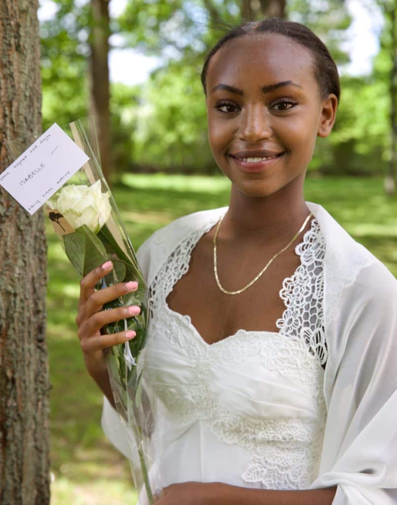 Festfotografen har her fotograferet en smilende kvindelig konfirmand. Hun står i nærheden af et træ og har en langstilket hvid rose i hånden